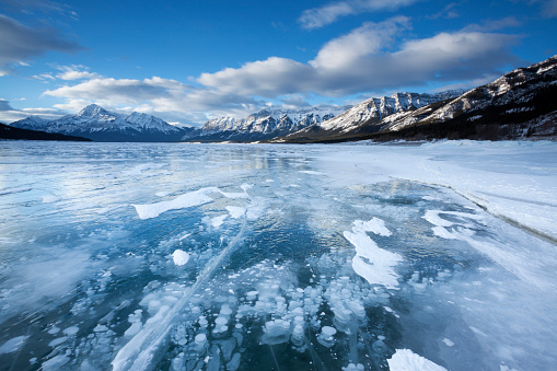 A fjord in Norway almost completely covered in ice with snow on top of it. The clouds in the sky block the sun. On the sides of the water are high mountains covered in snow.