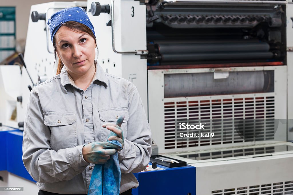 Woman working in printing plant wiping hands A serious woman working in a printing factory, looking at the camera.  She is standing next to a printing press, trying to wipe the blue ink off her hands with a rag. Cleaning Stock Photo