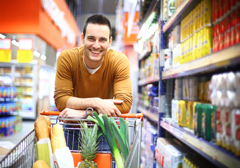 Smiling handsome in his late 20's leaning against shopping cart packed with groceries. He's in drinks aisle in local supermarket,wearing casual dark yellow sweater and facing camera. Aisle stretches behind him 