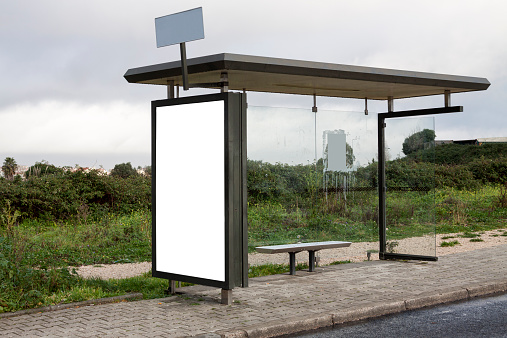 Bus stop in a rural setting with a large blank billboard on the side of the shelter. Grey, rainy day and vegetation in the background.