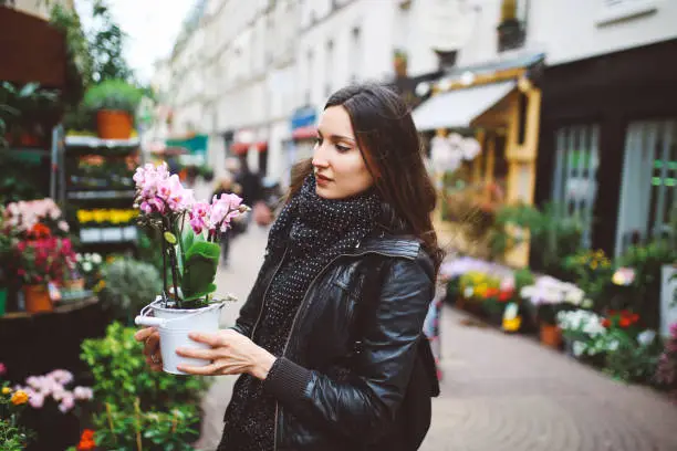 Vintage toned image of a young Parisian woman walking in the city center, buying flowers on a small street market. She is wearing a black leather jacket and a scarf, carrying a tote bag. Shot with wide angle large aperture prime lens for more shallow depth of field in motion.