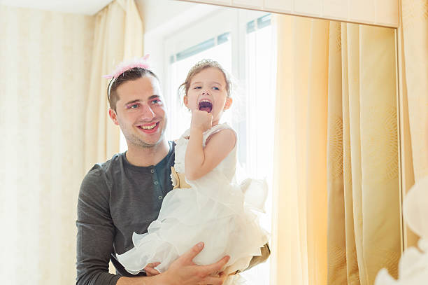 Cute little girl in princess dress that putt on colorful make up on her father, looking into a mirror