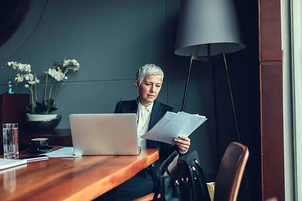 Mature businesswoman working, sitting in restaurant or office. In front of her, on the table, is laptop and in her hands are papers, examining documents. She is focused on her work.