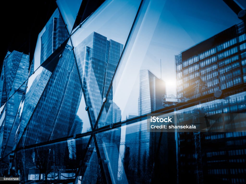 View of a modern glass skyscraper reflecting the buildings around View of a modern glass skyscraper reflecting the blue sky,blue toned image. Building Exterior Stock Photo