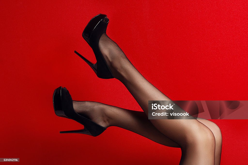 Slim female legs in dark stockings wearing high heels over Slim female legs in dark stockings wearing high heels . red background Sensuality Stock Photo