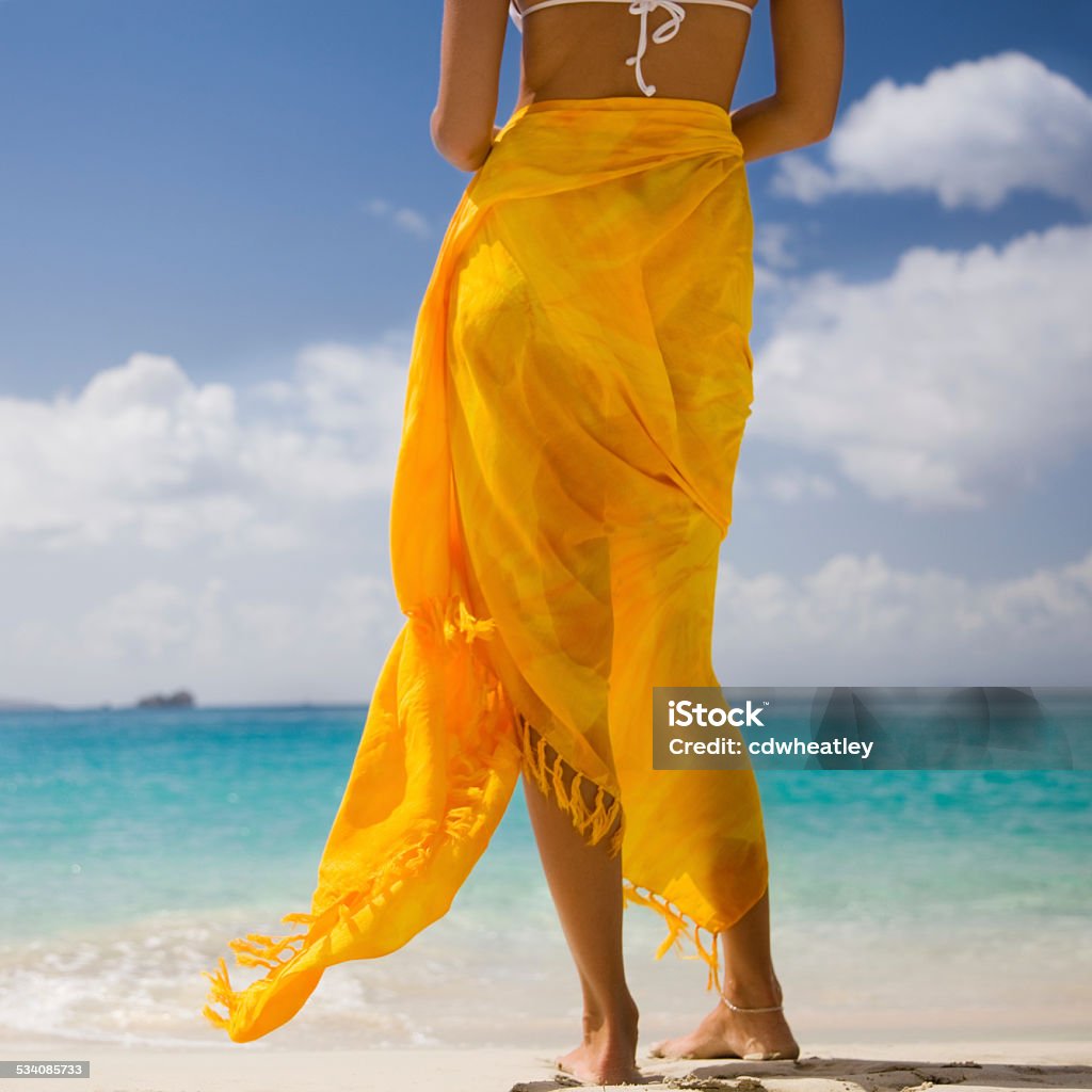 barefoot woman wrapped in sarong standing on a Caribbean beach unrecognizable and barefoot woman in white bikini wrapped in orange sarong standing on a beach in the Caribbean and enjoying the ocean view Sarong Stock Photo