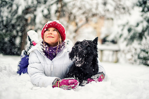 Happy little girl aged 9, is lying on her front on the snow hugging her black dog and looking up. She is smiling and admiring the beauty of the falling snow. Both the girl and the dog are snow covered after playing on the snow. The girl is wearing a white jacket and red cap.