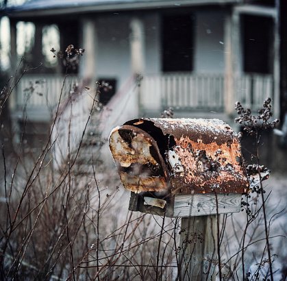 A long disused mail box at the end of the driveway of an abandoned cabin.  Light snow falling.