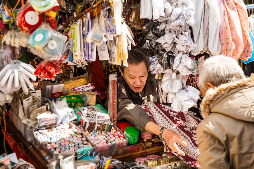 Kowloon, Hong Kong - January 9, 2010: chinese man in his market stall sells tailor equipments like needles,threads,clothes and zippers. He serves a man checking clothes.