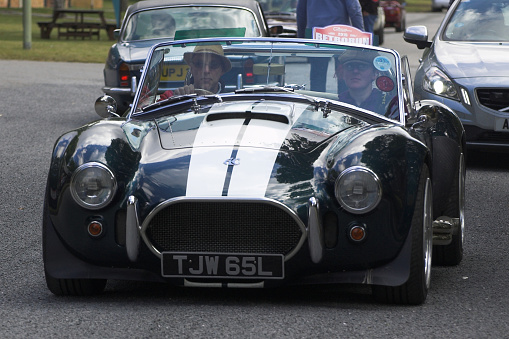 Bicester, Oxfordshire, England -  July 25th 2015: A black with white stripes AC Cobra replica kit car leaves a classic car show with two people onboard.