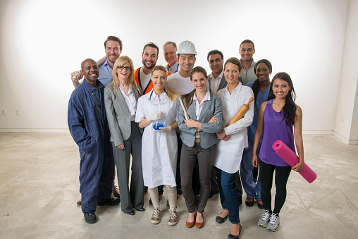 A multi-ethnic group of varied professionals are standing together in a group and are smiling while looking at the camera. There is a doctor, scientist, businesswoman, businessman, yoga instructor, baker, construction worker, teacher, mechanic, nurse, and a doctor.