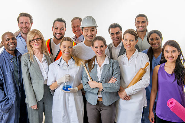 A multi-ethnic group of varied professionals are standing together in a group and are smiling while looking at the camera. There is a doctor, scientist, businesswoman, businessman, yoga instructor, baker, construction worker, teacher, mechanic, nurse, and a doctor.