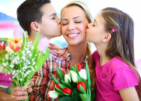 Boy and girl kissing their mom and giving her flowers. Mother is in her mid 30's.She's smiling and she has her eyes closed. Kids are aged from 9 to 11.All three of them wearing casual clothes.