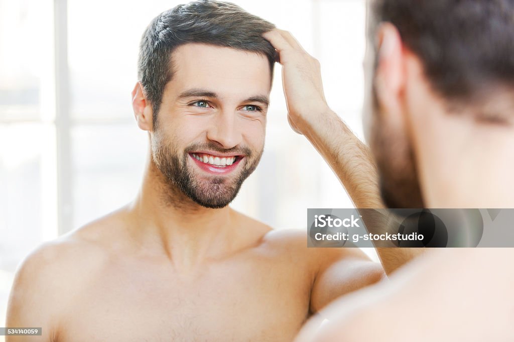 Starting new day with smile. Handsome young man touching his hair with hand and smiling while standing in front of the mirror Men Stock Photo