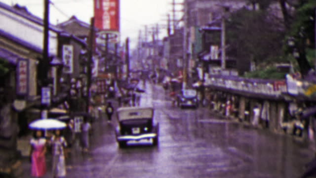 1951: Busy Japanese commercial street raining umbrellas drawn.