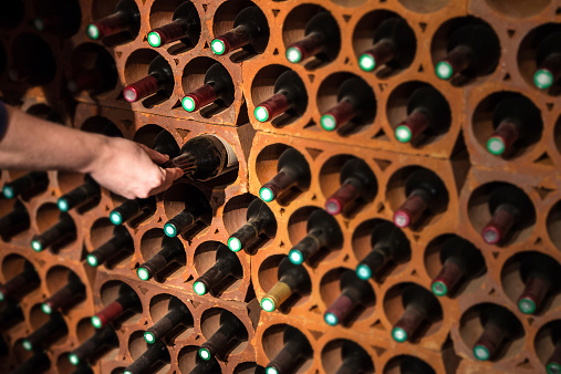 Selective focus of wine rack on wall in brick material full, to store red wine bottle in cellar, cool and in good conditions. A man is choosing one of those old wine bottle with dust to drink.