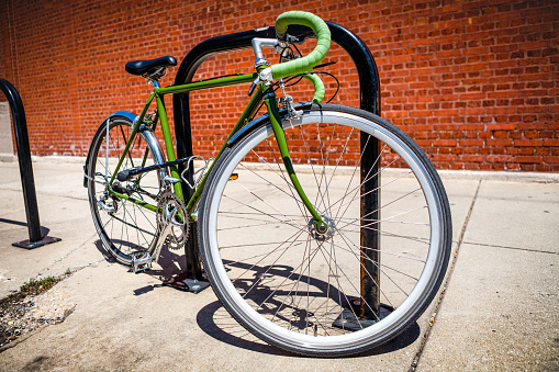 A green road bike with green handlebar tape and metal fenders locked to a staple bike rack on the sidewalk with red orange brick wall in the background.