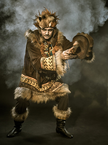 The mature man with appearance of Siberian indigenous people. The mustached man dressed ethnic clothing. He is dancing ritual dance and drumming in shaman drum. Studio shooting in smoke on black background. Sepia toned