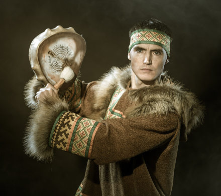 The young man with appearance of Siberian indigenous people. He is drumming in shaman drum. The attractive man dressed ethnic clothing looking at the camera. Studio shooting in smoke on black background. Sepia toned