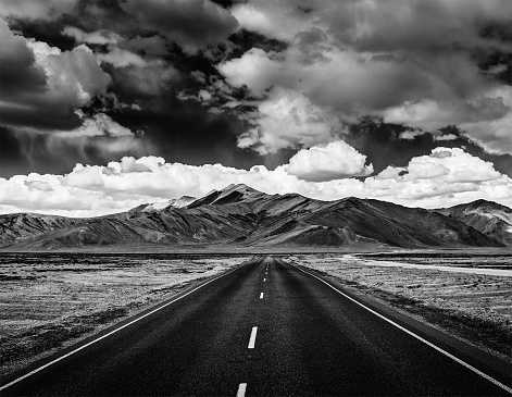 Travel forward concept background - road on plains in Himalayas with mountains and dramatic clouds. Manali-Leh road, Ladakh, Jammu and Kashmir, India. Black and white version
