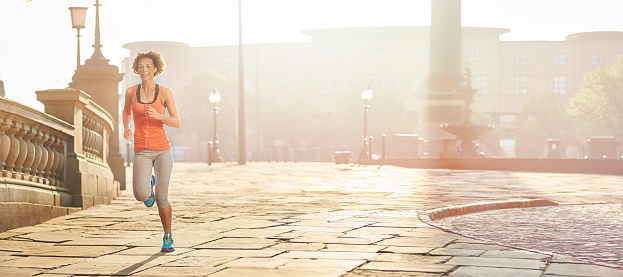 a young woman runs fin front of two sandstone historical buildings of liverpool , england in the morning. She is backlit by a rising low sun , and is pictured mid air ,  as she jogs across frame. She is wearing a vest top , trainers and leggings , and earphones. The image has a panoramic crop with plenty of copy space. The woman's hair is lifting as she takes her next stride .She is smiling and looks very healthy .