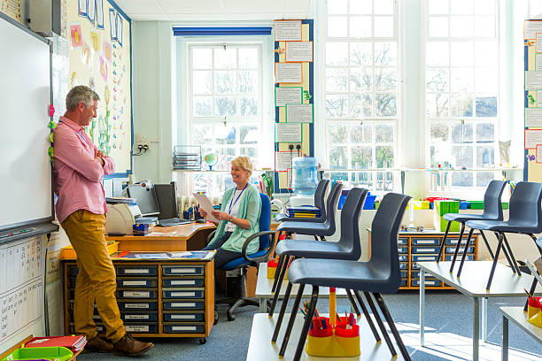 Teacher Staff Meeting Two teachers can be seen in a classroom after the children have left discussing school work. The meeting is casual and positive as both teachers appear happy. primary school assembly stock pictures, royalty-free photos & images
