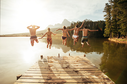 Portrait of young people jumping from pier into lake together. Friends jumping off the jetty at the lake on a sunny day.