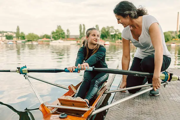 Single scull rowing. 12 years girl rowing single scull. She is preparing for new competitions. Rowing on Sava river. Coach giving instructions to girl rowing. They are looking at each other. Her mother is her coach.