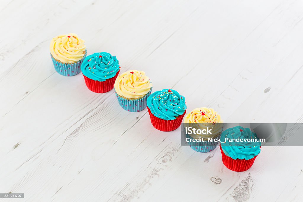 Tasty looking cupcakes aligned on bleached white wooden surface Selection of six tasty looking freshly baked cupcakes in vivid blue, red and butter cream colours diagonally aligned on white bleached pine timber surface. Indoors studio shot with Canon EOS. 2015 Stock Photo