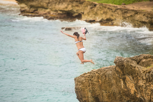 Young attractive ethnic female cliff jumping in tropical hawaii with snorkeling gear ready to go swimming with flippers and mask