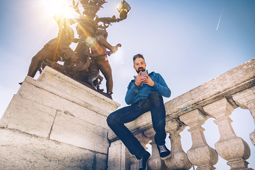 bearded tourist looking on mobile on Pont Alexandre in paris while sitting on balustrade