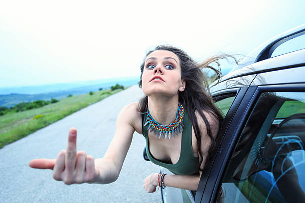 Young woman making rude gesture on the road Angry young woman showing trought the vehicle window and making rude gesture. Looking at camera. Road and nature on background. asshole stock pictures, royalty-free photos & images