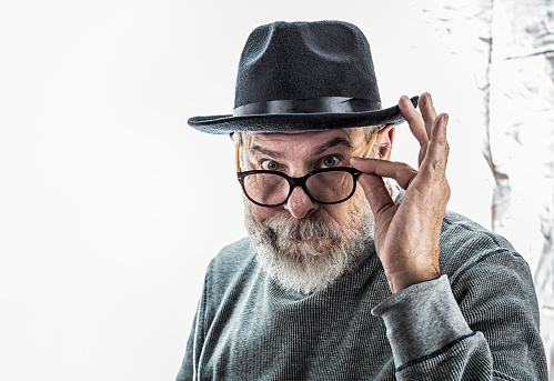 Quizzical, curious, dapper, senior adult man wearing a black fedora hat and a casual knit shirt is holding and looking over his black-rim glasses at the camera. Toned image.