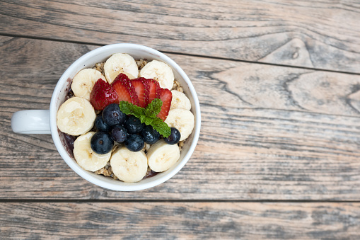 Very delicious acai bowls with fresh fruit strawberry, blueberry, banana granola and peppermint leaves on top in cute white cup on the wooden table. this smoothie dessert is good for summer in Hawaii.