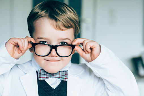 Boy wears black glasses, a bow tie and a lab coat. He adjusts his glasses and makes a funny face.