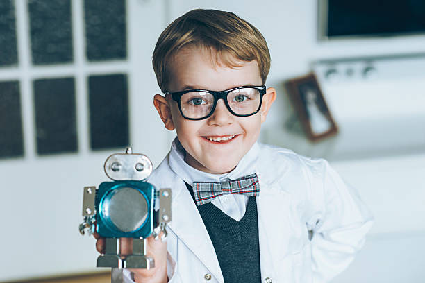 Boy dressed as a professor displays a toy robot. The boy wears a lab coat, glasses and a bowtie. The boy is proud and smiles while looking into the camera.