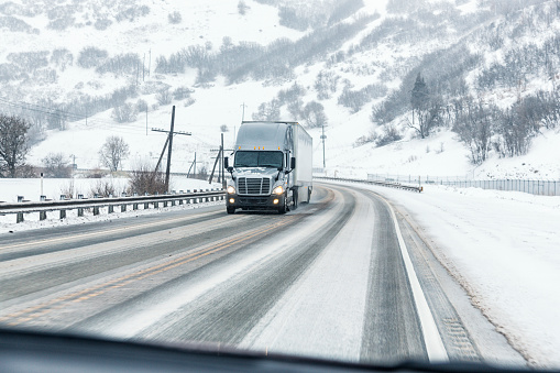 A semi-tractor trailer truck approaching during a snow storm - pulling hard around a sharp curve up a long hill - is about to pass a speeding car moving downhill on a frozen, slushy, slippery, Utah, USA winter mountain highway.