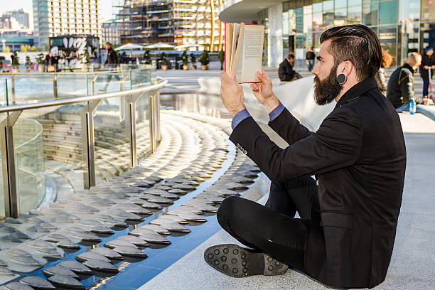 Young man (hipster style) with big earring and full beard reading a book outdoor in a modern square