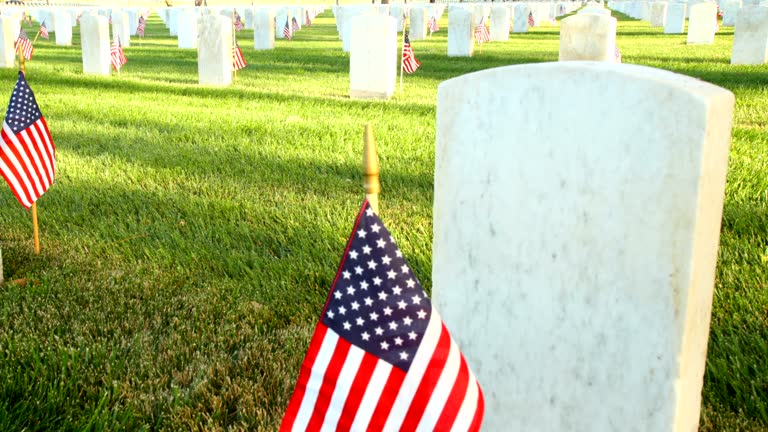 Dolly camera moves up past a marble tombstone at a US National Cemetery. Many more tombstones can be seen in the background. Small flags are placed next to each tombstone.
