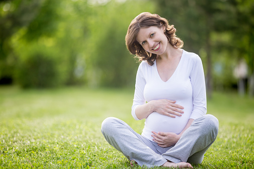 Portrait of happy young pregnant model sitting with crossed legs on grass lawn and looking at camera. Smiling future mom expecting baby caressing her belly. Maternity concept. Copy space
