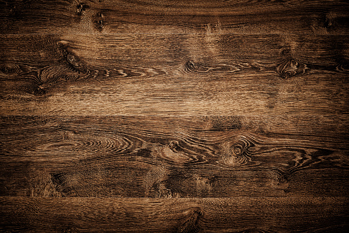 Overhead view of dark brown floor texture. A wood background with multiple planks placed close together.  The planks feature a variety of light and dark brown shades.  The darkest shades are on the corners, the lightest on the middle. There are variety of knots and swirls on it.