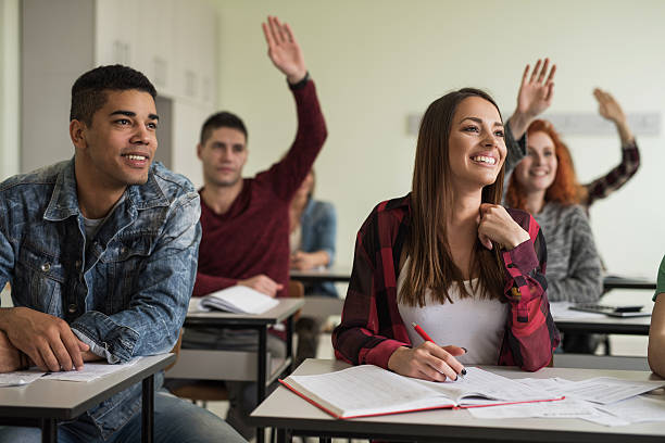 Smiling female student with her classmates sitting in the classroom. Group of high school students listening a lecture in the classroom. Focus is on happy female student. hand raised classroom student high school student stock pictures, royalty-free photos & images