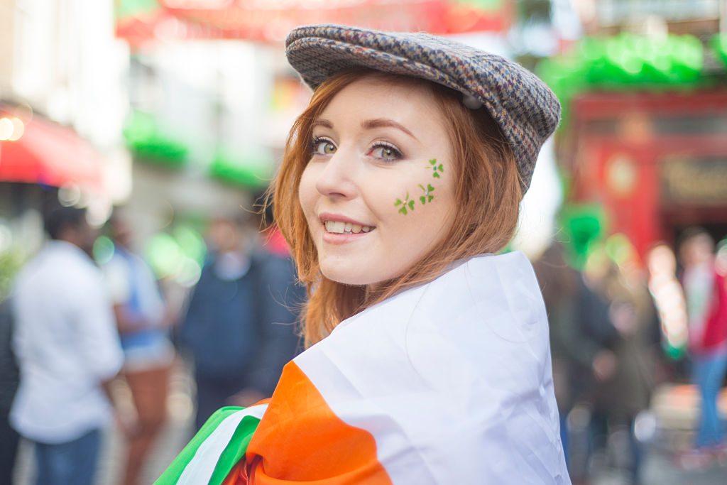 Smiling pretty red haired Irish girl with tweed hat, wrapped in the Irish flag and shamrocks painted on her face, Temple Bar, Dublin, Ireland. 