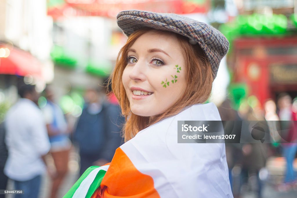 Beautiful Irish girl on St. Patricks Day, Dublin, Ireland. Smiling pretty red haired Irish girl with tweed hat, wrapped in the Irish flag and shamrocks painted on her face, Temple Bar, Dublin, Ireland.  St. Patrick's Day Stock Photo