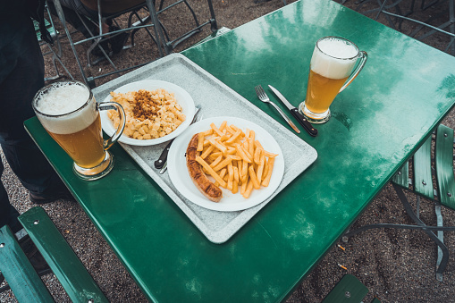 High Angle View of Fast Food Meals of Pasta, French Frieds and Sausage on White Plates and Served on Cafeteria Tray with Mugs of Light Beer on Green Table in Outdoor Dining Area