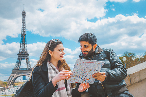 Happy young tourist couple holding and reading paper citymap, looking around together in Paris, France against Eiffel Tower from Trocadéro