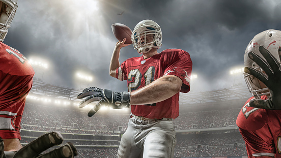 A close up low angle view of American football player holding football and about to throw whilst being protected by teammates. Action takes place in generic outdoor floodlit stadium full of spectators under stormy evening sky. Players are wearing generic kit. Image created with intentional lens flare and lighting effects.
