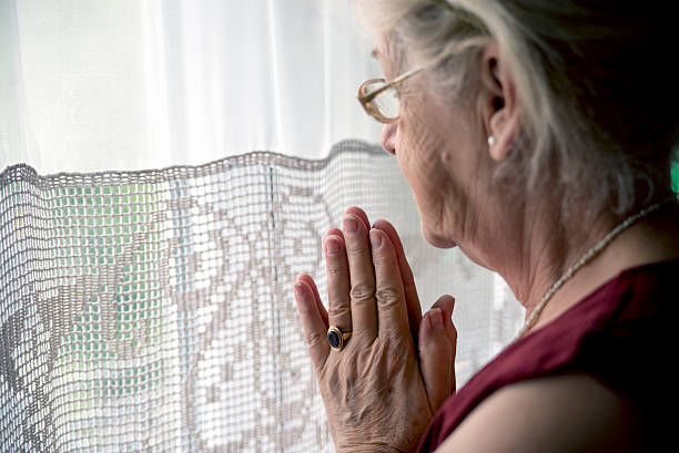 Senior woman lost in thought and praying An elderly lady sitting near the window in the kitchen, Lovely grandmother looking in a window and praying grandmother real people front view head and shoulders stock pictures, royalty-free photos & images