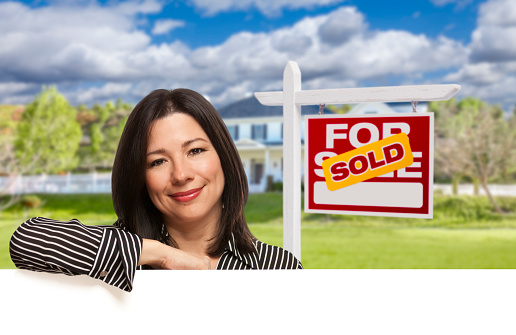 Pretty Hispanic Woman Leaning on White in Front of Beautiful House and Sold For Sale Real Estate Sign.