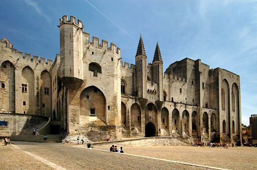 Avignon, France - June 16, 2005: Facade of the Palace of the Popes (Palais des Papes) in France. In front of the photo is a square with almost no people. Some persons are sitting on a somewhat higher part of the square.